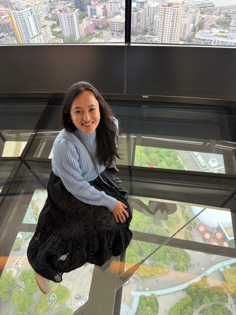 A woman sitting on a glass floor observation deck of the Space Needle in Seattle, smiling, with a city skyline visible below.