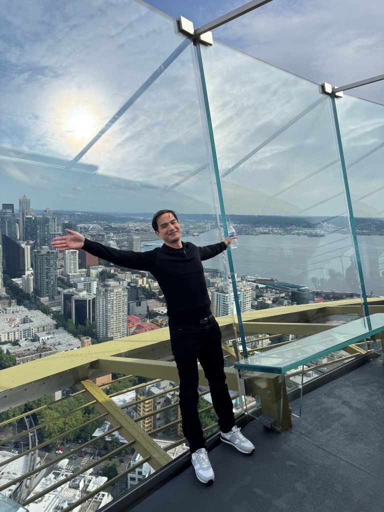 A person standing with arms outstretched on the observation deck of the Space Needle in Seattle, with a panoramic view of the city skyline and waterfront in the background.