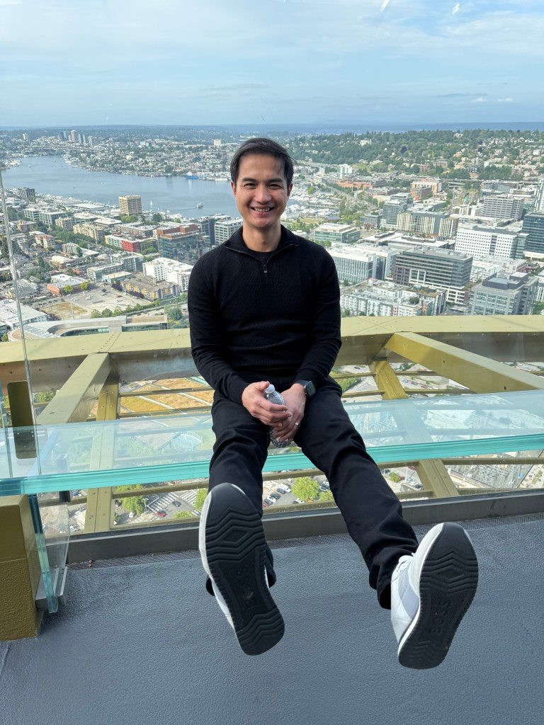 A person sitting on a glass-paneled observation deck at the Space Needle in Seattle, with a panoramic view of the cityscape and Elliott Bay in the background.
