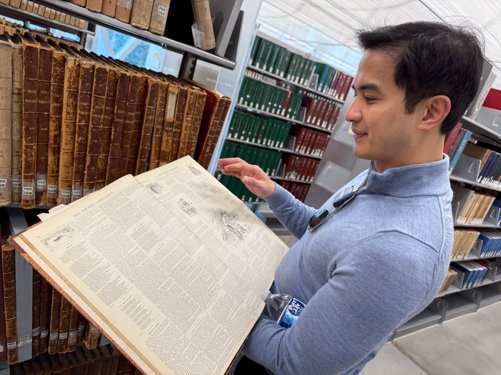 A person examining an open historical book in a library, surrounded by shelves filled with old leather-bound volumes.