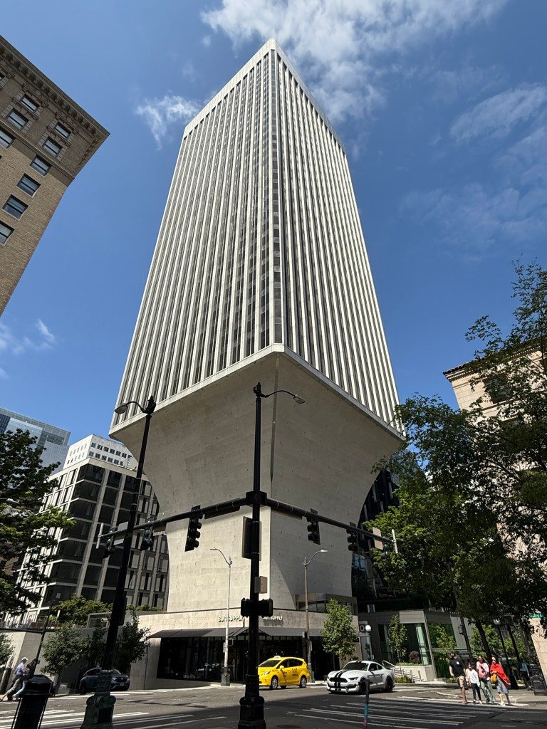 A tall, modern skyscraper with a unique architectural design, featuring a wider base and a slender upper portion, set against a clear blue sky in Seattle.